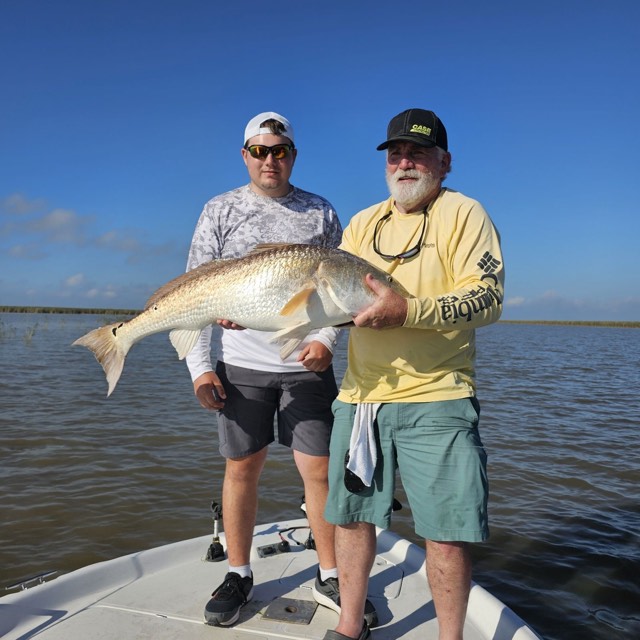 Two men on a boat hold a large redfish in shallow water. Clear, blue sky.