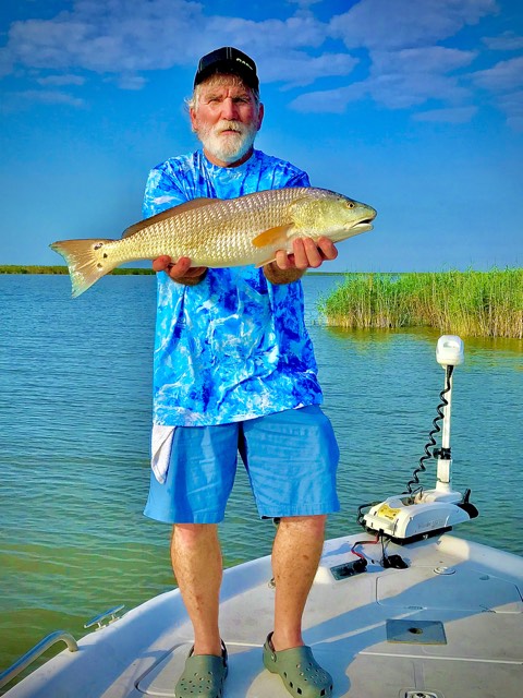 Man holding a redfish on a boat, blue sky, water, and marsh in the background.