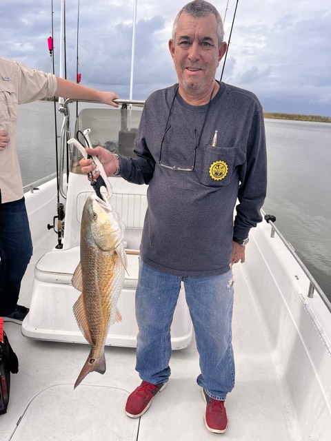 Man on boat holding up a fish, wearing a long sleeve shirt and jeans. Water and shoreline visible.