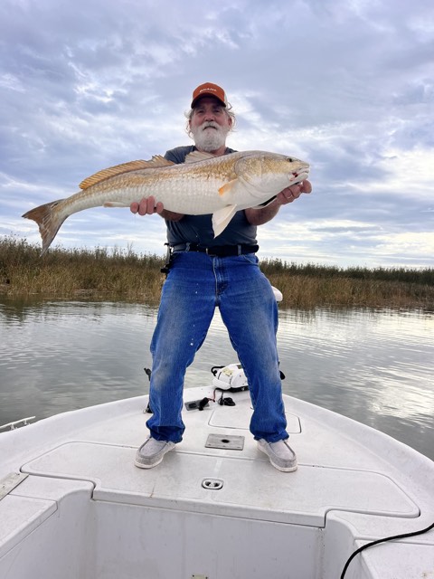 Man holding a large redfish on a boat, cloudy sky, marsh background.