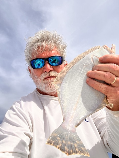 Man holding a flat fish, possibly a flounder, with a light-colored body. He wears sunglasses against a cloudy sky.