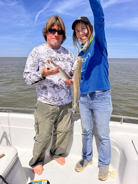 Two people fishing from a boat. One holds a fish up, the other holds one. Sunny day, water background.
