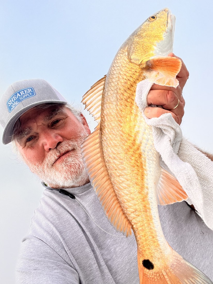 Man holding up a reddish-orange redfish with a single black spot near its tail; outdoors.