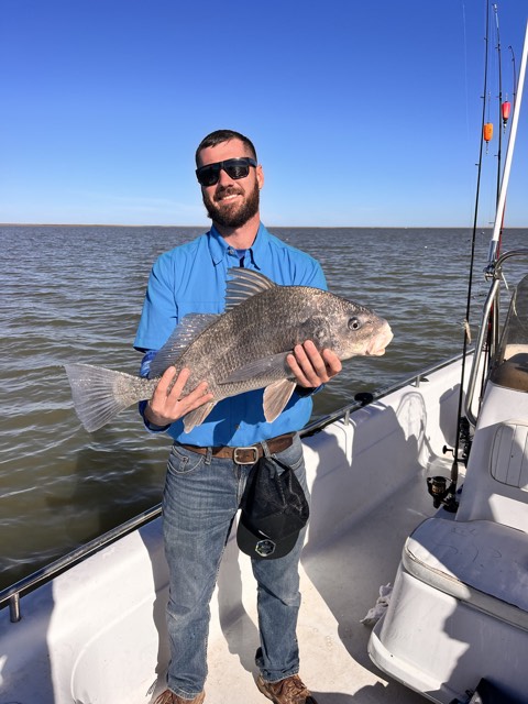 Man holding a large black drum fish on a boat, wearing sunglasses and a blue shirt, smiling at camera.