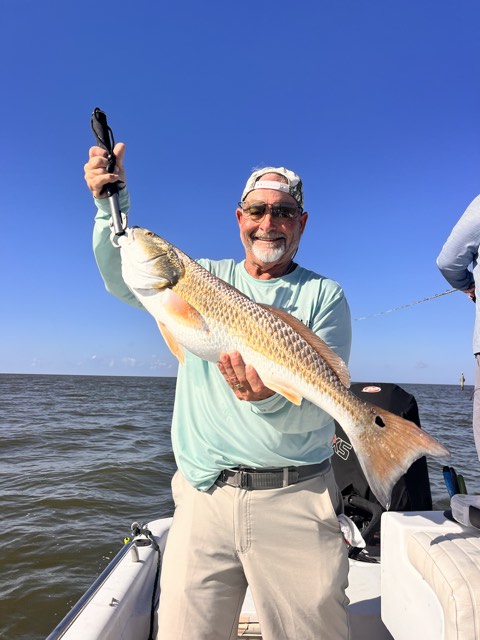 Man on boat holding up a redfish with a fishing scale. Blue sky and water in the background.