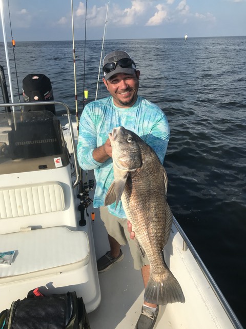 Man on a boat holding a large black fish, smiling. Sunny day, open water.