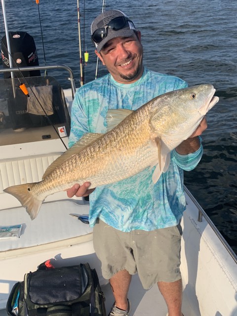 Man on a boat holding a large redfish he caught, smiling. Blue water and fishing rods visible.